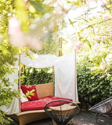 Gemütlicher Sitzplatz im Garten mit Sitzbank, rotem Kissen, kleinem Tisch und Stühlen, umgeben von grünen Pflanzen.