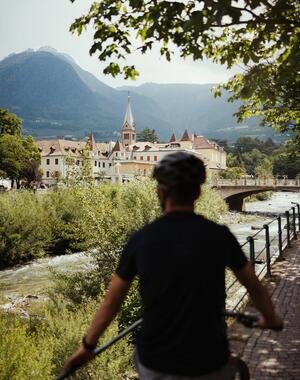 Mann auf dem Fahrrad auf der Promenade in Meran mit Blick auf einen Fluss, die Stadt und die Berge.