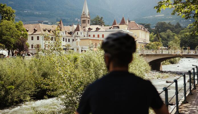 Uomo in bicicletta sulla passeggiata di Merano con vista su un fiume, sulla città e sulle montagne.
