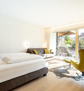 Helles Schlafzimmer mit Doppelbett, Sessel, Sofa, Holzboden und Zugang zum Balkon mit Blick ins Grüne.