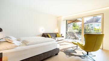 Helles Schlafzimmer mit Doppelbett, Sessel, Sofa, Holzboden und Zugang zum Balkon mit Blick ins Grüne.