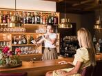 Female bartender behind the counter of a wood-panelled bar, with a woman seated in front.
