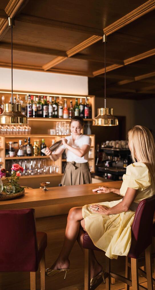 Barkeeperin  hinter dem Tresen einer Bar mit Holzverkleidung, davor sitzt eine Frau.