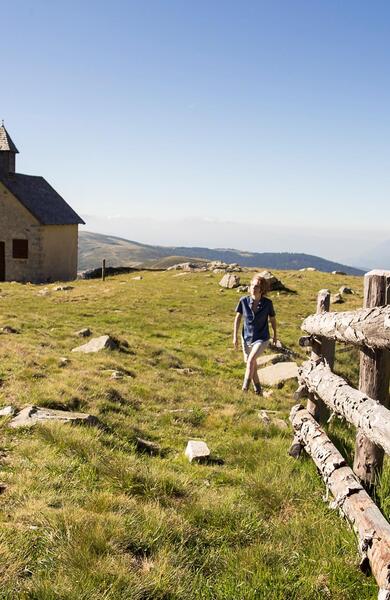 Wanderer auf einem Bergpfad neben einem Holzzaun, mit einer kleinen Kapelle und Bergen im Hintergrund.