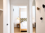 Flur mit Holzboden, ovalem Wandspiegel und Garderobenhaken, mit Blick ins Schlafzimmer.