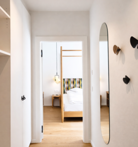 Flur mit Holzboden, ovalem Wandspiegel und Garderobenhaken, mit Blick ins Schlafzimmer.