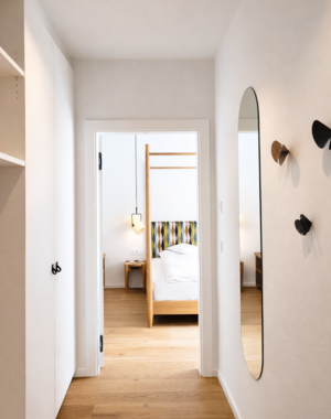 Corridoio con pavimento in legno, specchio ovale a parete e ganci appendiabiti, con vista sulla camera da letto.