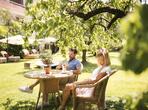 Couple sitting at a table in a green garden, surrounded by lawn, trees, loungers and parasols.
