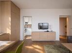 Wohnbereich mit grauem Sofa, grünem Polstersessel, Fernseher auf einem Sideboard und Blick in die Küche.