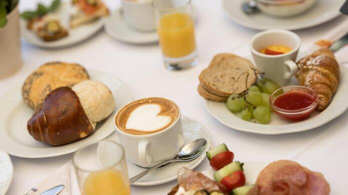 Tavola apparecchiata per la colazione con cappuccino, succo d’arancia e pietanze salate e dolci.