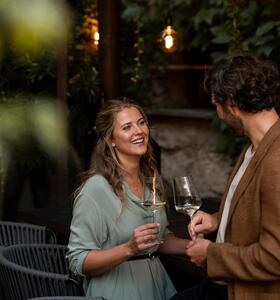 Una coppia con calici di vino in un giardino, circondata da piante e lampade sospese.