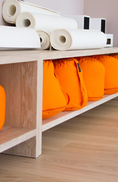 Orange meditation cushions and rolled-up white yoga mats on a shelf in a bright room.