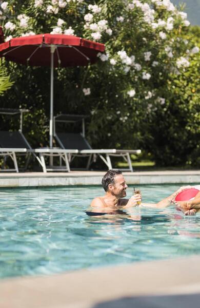 Mann und Frau mit Getränken im Außenpool, die Frau auf einem rot-weißen Schwimmring, mit Liegen und roten Sonnenschirmen im Hintergrund.