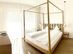 Helles Schlafzimmer mit Doppelbett, Vorhängen, Holzboden und Blick auf den Balkon.