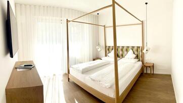 Helles Schlafzimmer mit Doppelbett, Vorhängen, Holzboden und Blick auf den Balkon.
