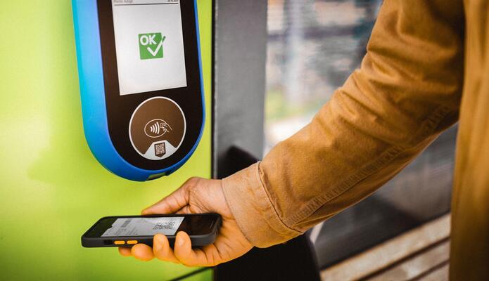 Eine Hand hält ein Smartphone mit einem QR-Code auf dem Display unter ein Ticketlesegerät.