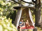 Couple sitting on a covered garden lounger on a green lawn, surrounded by trees and plants.