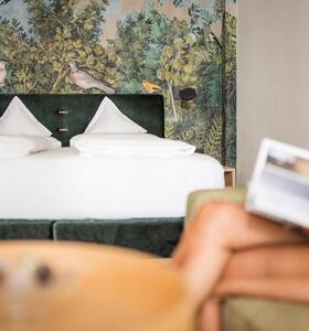Donna legge su un divano verde davanti a un letto matrimoniale con carta da parati botanica.