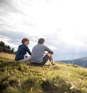 Zwei Wanderer sitzen auf einer Almwiese mit Blick auf die umliegende Berglandschaft.