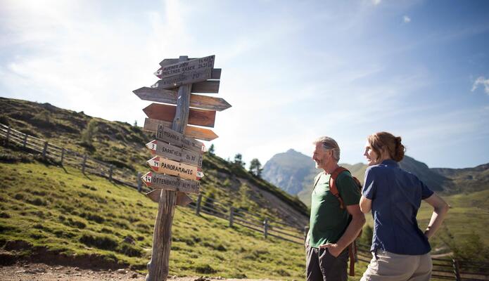 Due escursionisti su un sentiero di montagna accanto a un cartello segnavia, con pascoli alpini ondulati e montagne sullo sfondo.