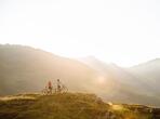 Due ciclisti su un sentiero di montagna, con montagne sullo sfondo e prati soleggiati in primo piano.
