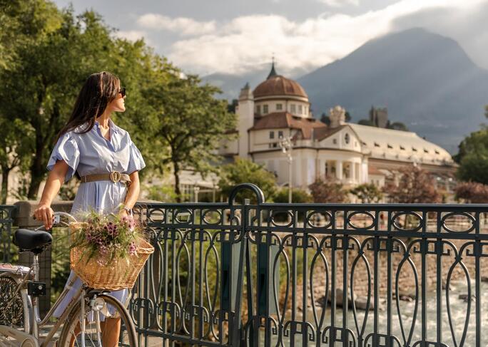Una donna in abito estivo attraversa un ponte spingendo una bicicletta, con il Kurhaus, alberi e montagne boscose sullo sfondo.