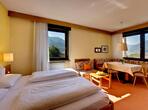 Zimmer mit Doppelbett, einer Sitzecke mit Tisch, Bank und Stühlen, davor Fenster mit Blick in die umliegende Landschaft.