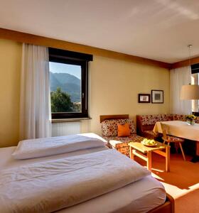 Zimmer mit Doppelbett, einer Sitzecke mit Tisch, Bank und Stühlen, davor Fenster mit Blick in die umliegende Landschaft.