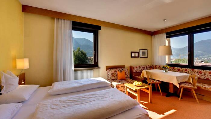 Zimmer mit Doppelbett, einer Sitzecke mit Tisch, Bank und Stühlen, davor Fenster mit Blick in die umliegende Landschaft.