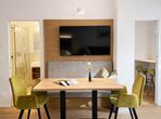 Essbereich mit Holztisch, grünen Polsterstühlen, Sitzbank und Wandfernseher.