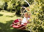 Woman sitting in a hanging chair, surrounded by green lawn, trees and plants.