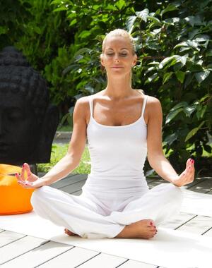 Woman dressed in white meditating cross-legged on a white yoga mat, surrounded by green plants.
