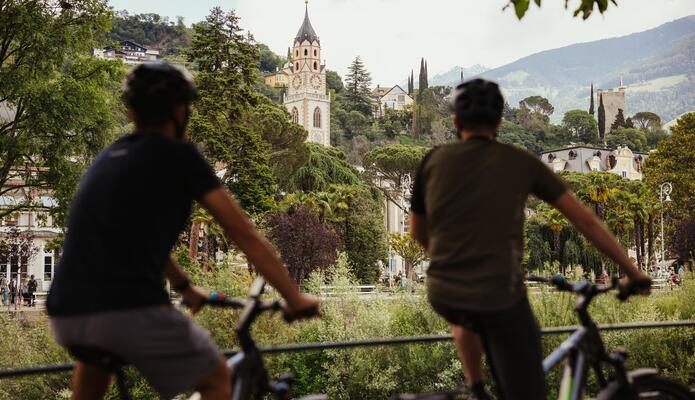 Zwei Fahrradfahrer auf der Promenade in Meran mit Blick auf die Stadt und die umliegenden Berge.