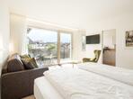 Helles Schlafzimmer mit Doppelbett, Sessel, Sofa, Holzboden mit Blick in die Küche und Zugang zum Balkon mit Blick ins Grüne.