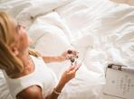 Woman sitting on a double bed with white bedding, holding a small plate of chocolates and a magazine.