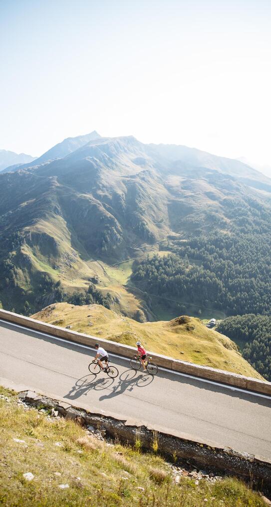 Zwei Fahrradfahrer auf einer Asphaltstraße im Hintergrund hügelige Wiesen und Wälder im Sonnenlicht.
