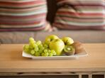Primo piano di un piatto di frutta con uva, mele e kiwi su un tavolino in legno davanti al divano.