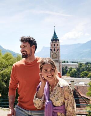 A couple stands on the Tappeiner Promenade in Merano, surrounded by trees and plants, with the parish church and a hilly landscape in the background.