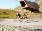 Due ciclisti su una strada di montagna davanti a una struttura in legno dalle forme angolari, con prati e montagne sullo sfondo.