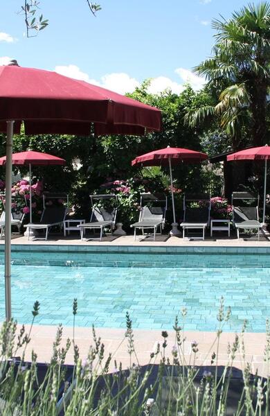 Piscina all’aperto con lettini e ombrelloni rossi, circondata da alberi e piante.
