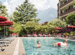 A man and a woman with an inflatable ring in the outdoor pool, surrounded by sun loungers, red parasols and plants.