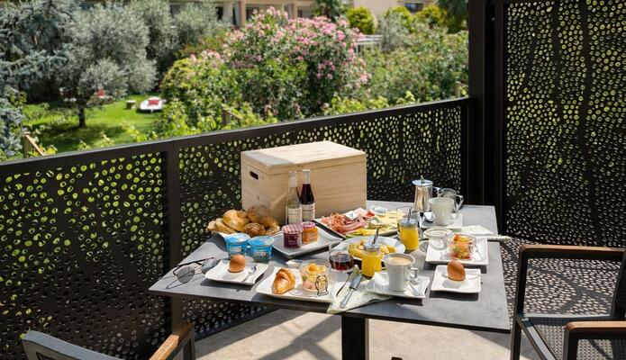 Tavolo da colazione apparecchiato su una terrazza con diversi piatti e bevande, circondato da piante e con vista sul verde.