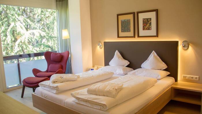 Camera nell’appartamento Champagner con letto matrimoniale, poltrona rossa e ampia vetrata con vista sul verde.