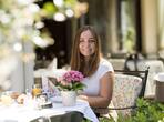 A woman sits at a table set for breakfast on a terrace and smiles at the camera.