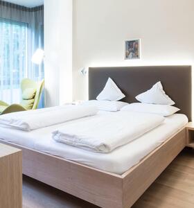 Deluxe Suite mit Doppelbett und gepolstertem Kopfteil, grünem Sessel und Stehlampe neben der Fensterfront.