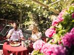 Couple sitting at a set table on a sunny garden terrace, surrounded by flowers and green plants.