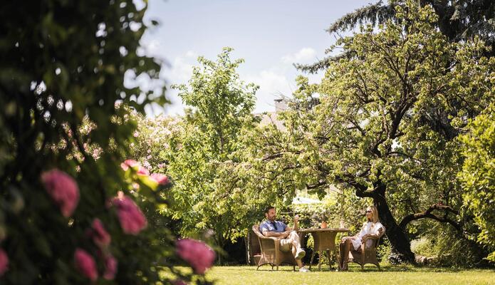 Coppia seduta a un tavolo in un giardino verde, circondata da prato e alberi.
