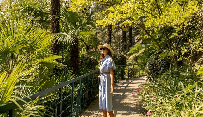 Una donna con cappello di paglia e occhiali da sole si trova sulla passeggiata Tappeiner a Merano, circondata da palme e piante in fiore.