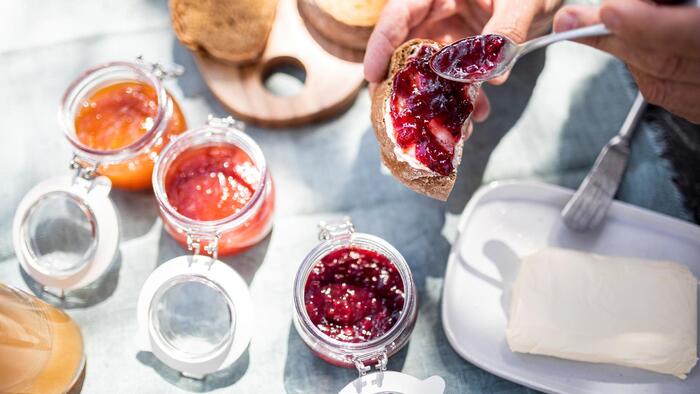 Close-up of hands spreading red jam on a buttered slice of bread, next to a block of butter and open jars of jam.