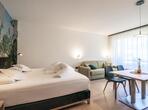 Doppelzimmer mit Doppelbett, Sofa und Sitzbereich mit Holztisch und gepolsterten Stühlen vor einer großen Fensterfront.